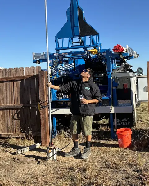A tech holds a length of pipe and looks up at the raised blue pump hoist mast under a clear blue sky.