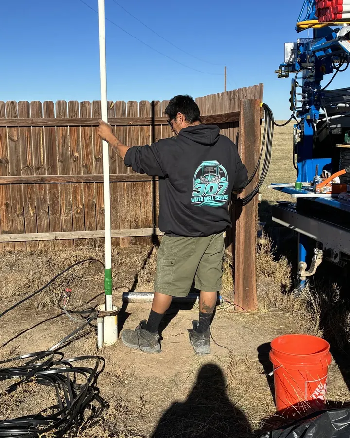 A 307 Water Well Service tech in a branded hoodie guides white pipe down into a well casing, with the pump hoist truck alongside on dry prairie.